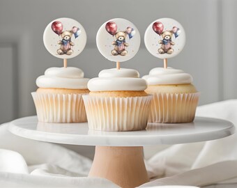 May include: Three cupcakes with white frosting and decorative toppers featuring a teddy bear holding balloons. The cupcakes sit on a white marble cake stand with a light pink base, against a soft, neutral background.