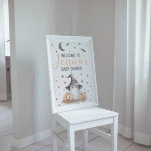 K&ouml;nnte beinhalten: Ein wei&szlig;es Schild mit schwarzem Rahmen und einer Babyparty-Ank&uuml;ndigung zum Thema Halloween. Das Schild lautet "Willkommen zur Babyparty von Jessica" und zeigt einen Geist, der einen Hexenhut tr&auml;gt und zwei K&uuml;rbisse h&auml;lt.