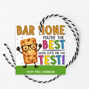 May include: A gift tag with a cartoon granola bar wearing glasses and arms up. Text reads "BAR NONE YOU'RE THE BEST GOOD LUCK ON THE TEST!" with "FROM: MRS. ROBINSON" at the bottom. The tag is white with a green border and a black and white string.