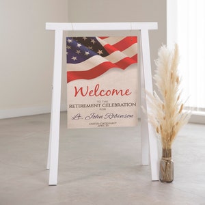 May include: A white easel sign with a welcome sign for a retirement celebration for Lt. John Robinson, United States Navy, April 20. The sign features an American flag with red, white, and blue colors.