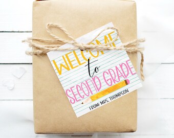 May include: A gift tag with a blue and white striped background and the text "Welcome to Second Grade" in yellow and black lettering. A yellow pencil with a pink eraser is drawn on the tag. The text "From: Mrs. Thompson" is printed in black at the bottom of the tag.