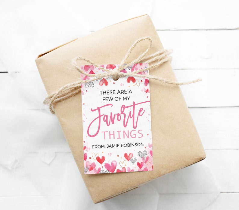 K&ouml;nnte beinhalten: Ein wei&szlig;er Geschenkanh&auml;nger mit rosa Herzen und goldenem Glitzer. Der Anh&auml;nger lautet "Das sind ein paar meiner Lieblingsdinge." und "Von: Jamie Robinson".