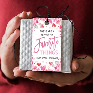 K&ouml;nnte beinhalten: Ein wei&szlig;er Geschenkanh&auml;nger mit rosa und wei&szlig;en Herzen und goldenem Glitzer. Der Anh&auml;nger lautet "Das sind ein paar meiner Lieblingssachen. Von Jamie Robinson."