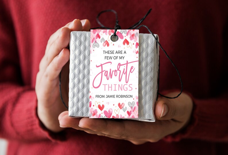 K&ouml;nnte beinhalten: Ein wei&szlig;er Geschenkanh&auml;nger mit rosa und wei&szlig;en Herzen und goldenem Glitzer. Der Anh&auml;nger lautet "Das sind ein paar meiner Lieblingssachen. Von Jamie Robinson."