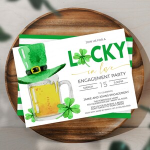 May include: Watercolor engagement party invitation with a green leprechaun hat, beer mug, and shamrocks. The invitation reads "Lucky in Love" with event details, set on a wooden tray.