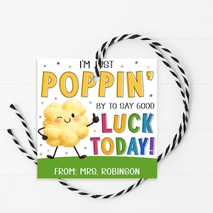 Puede incluir: Una etiqueta de regalo cuadrada con un personaje de palomitas de dibujos animados y el texto "I'm just poppin' by to say good luck today!" en amarillo, verde, azul y rosa. La etiqueta tiene una pancarta verde en la parte inferior con el texto "From: Mrs. Robinson". Una cuerda en blanco y negro está adjunta.