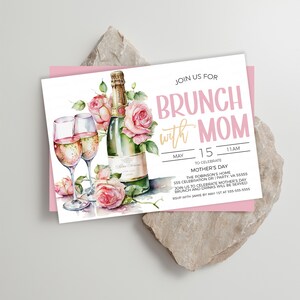 May include: A white invitation card with pink accents. The card features a watercolor illustration of champagne flutes and a bottle of champagne with pink roses. The text on the card reads "Join us for Brunch with Mom" and includes details for a Mother's Day celebration.