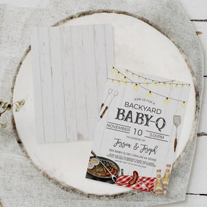 May include: A white woodgrain background with a white invitation card. The invitation card has a rustic design with string lights, a spatula, and a grill. The text on the card reads "Join us for a Backyard Baby-Q, November 10, 5:00 PM, In honor of Jessua & Joseph, 555 Baby-Q Dr. Grillin VA 55555, Join us to celebrate a baby shower, Food, drinks, bring a pack of diapers, RSVP by Nov 1st at 555-555-5555".