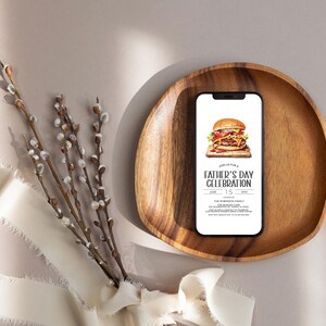 May include: A smartphone mockup with a Father's Day celebration invitation on the screen. The invitation features a burger and the text "Join us for a Father's Day Celebration" with the date and time. The phone is on a wooden plate with a white ribbon and a sprig of pussy willow branches in the background.