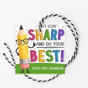 Puede incluir: Una etiqueta de regalo cuadrada con un lápiz de dibujos animados con gafas y el texto "JUST STAY SHARP AND DO YOUR BEST!" en letras coloridas. La etiqueta tiene una base verde y está atada con una cuerda en blanco y negro. La etiqueta dice "FROM: MRS. ROBINSON".