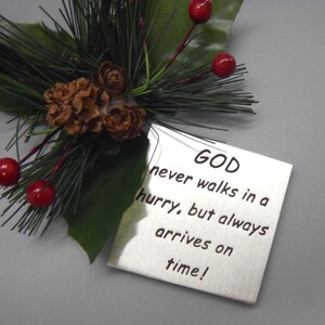 May include: A silver metal ornament with a quote that reads "GOD never walks in a hurry, but always arrives on time!" The ornament is decorated with a pine sprig and red berries.