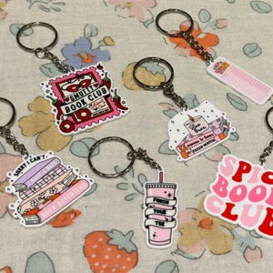 May include: Six pink and red keychains with different book club themes. The keychains feature phrases like "Smutty Book Club", "Spicy Book Club", "Sorry I Can't, I Have a Book Boyfriend", and "Finish Your Wine".