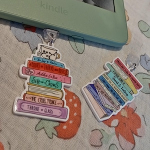 May include: Two book-themed earrings, one with a teacup design, and titles including "Court of Thorns and Roses" and "Throne of Glass." They rest on a floral fabric, near a Kindle e-reader.
