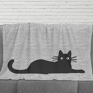 May include: A gray and white patterned throw blanket with a black silhouette of a cat lying down. The cat has large, round eyes and a long tail.