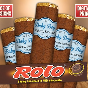 May include: Four brown chocolate bar wrappers with blue and white labels that say "It's a Baby Boy" with a name and date. The wrappers are arranged in a row with the chocolate bar in the front. The chocolate bar is a Rolo with the text "Chewy Caramels in Milk Chocolate" printed on the wrapper.