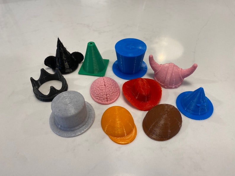 Puede incluir: Una colecci&oacute;n de diez sombreros impresos en 3D en varios colores y estilos, incluyendo una corona, un sombrero de copa, un casco vikingo, un sombrero c&oacute;nico y un sombrero con forma de cerebro.