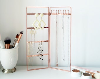 May include: A rose gold jewelry stand with multiple hooks for necklaces and earring posts. The stand is folded open and has several necklaces and earrings hanging from it.