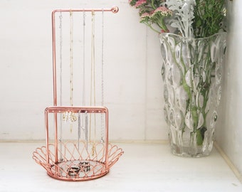 May include: Rose gold jewelry stand with a tiered design. The stand has a round top with a bar for hanging necklaces and a lower tier with a wire basket for rings. The stand is empty.