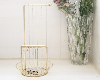 May include: Gold metal jewelry stand with a tiered design. The top tier has a bar for hanging necklaces and the bottom tier has a square tray with a mirror for storing earrings and rings. The stand is on a white surface.