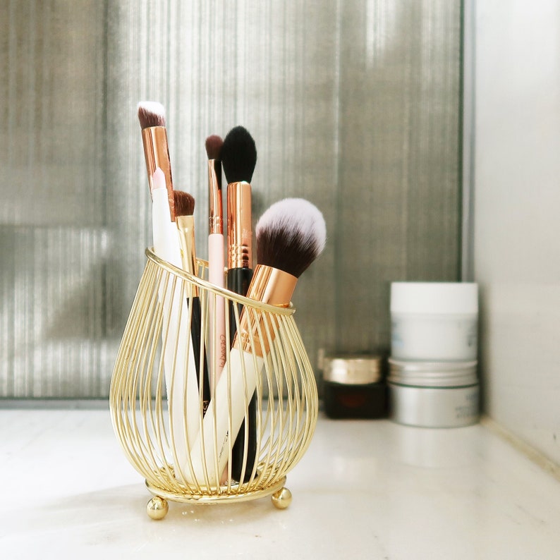 May include: A gold metal makeup brush holder with a pearlescent white makeup brush, a black makeup brush, and a pink makeup brush.