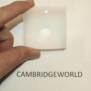 May include: A white, square, slightly curved piece of glass. The glass is translucent and has a slight sheen. The glass is being held by a hand.