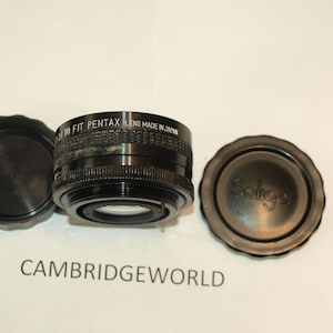 Puede incluir: Un adaptador de lente de cámara negro con el texto "TO FIT PENTAX LENS MADE IN JAPAN" impreso en el lateral. El adaptador viene acompañado de dos tapas de lente negras, una de las cuales tiene la palabra "Soligor" impresa.