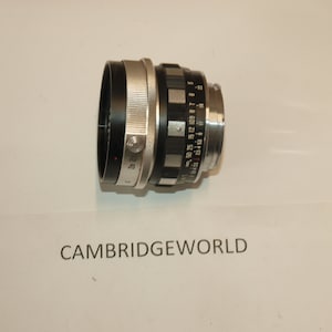 Può includere: Un obiettivo per fotocamera argentato e nero con una scala di profondità di campo e un tappo dell'obiettivo nero.
