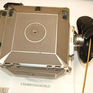 Puede incluir: Una cámara vintage plateada y marrón con un fuelle negro adjunto. La cámara tiene una lente circular y el texto "Leica" y "Made in Germany" en el lateral.