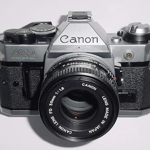 May include: A silver Canon AE-1 Program film camera with a Canon FD 50mm 1:1.8 lens attached. The lens is black and has the text "Canon Lens FD 50mm 1:1.8 Canon Lens Made in Japan" printed on it.