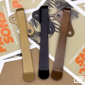 May include: Three metal pocket clips in gold, black, and brown. Each clip has a curved design with a hole at the top for attachment. The background features cards with the text "PACIFIC SONS AMERICAN SUPPLY COMPANY".