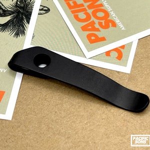 Puede incluir: Llavero de metal negro con un agujero en el centro. El llavero es curvo y tiene un extremo redondeado. El llavero está sobre una superficie marrón.