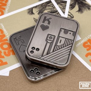May include: Two metallic playing card-shaped objects, one on top of the other, with a King of Hearts design. The top card features the letter "K" and a heart symbol. The background includes cards with the text "PACIFIC SONS".