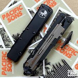 Puede incluir: Dos cuchillos automáticos negros, uno abierto y otro cerrado, se muestran sobre un fondo de tarjetas. Las tarjetas presentan el texto "PACIFIC SONS AMERICAN SUPPLY COMPANY" en naranja y negro. El cuchillo abierto revela un mecanismo interno plateado.