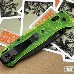 May include: A neon green and black folding knife with a textured handle and a black pocket clip. The knife is resting on a pile of cards with a palm tree design and the words "PACIFIC SONS".