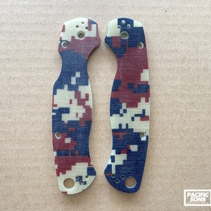May include: A pair of red, white, and blue camouflage patterned knife scales. The scales are made of a textured material and have a slightly rounded shape.