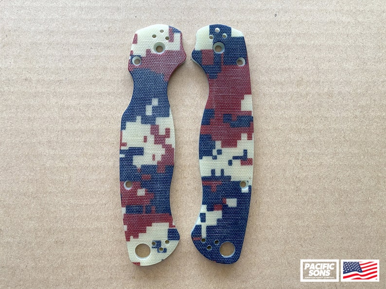 May include: A pair of red, white, and blue camouflage patterned knife scales. The scales are made of a textured material and have a slightly rounded shape.