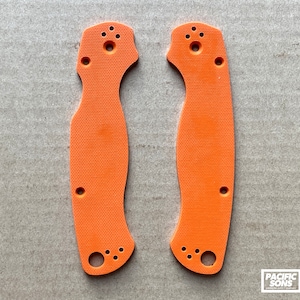 May include: Two orange plastic knife scales with holes for mounting hardware. The scales are textured and have a slightly curved shape.