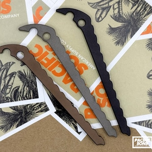 May include: Three metal tent stakes in matte brown, grey, and black finishes. Each stake features a curved hook top with a circular hole and a serrated edge along one side for secure ground anchoring. The stakes are arranged diagonally.