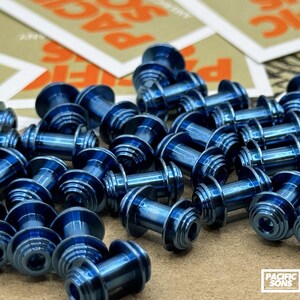 May include: A close-up shot of a pile of small, metallic blue objects. The objects have a cylindrical body with a flared, ridged head and base. The objects are scattered on a light brown surface, with some stickers in the background.