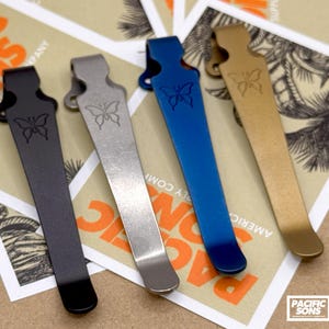 May include: Four metal pocket clips in black, silver, blue, and gold, each with a butterfly design. They are displayed on a background of tan and orange paper with the text "PACIFIC SONS" and an American flag.