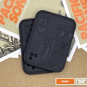 May include: Two rectangular black fidget tools with rounded corners, featuring a debossed design of a playing card king of hearts. The surface includes raised lines, a heart symbol, the letter K, and three small circular indentations in the corner.
