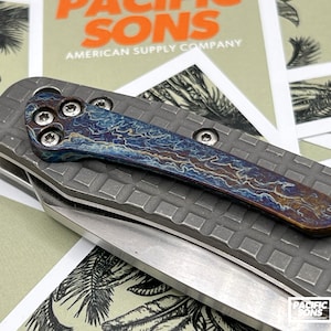 Op de afbeelding: Een close-up van een blauw en gouden titanium zakmes met een getextureerd handvat en een gedeeltelijk geopend mes. Het mes ligt op een stuk papier met de tekst "Pacific Sons American Supply Company" erop gedrukt.