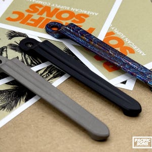 May include: Three metal bottle openers arranged diagonally. One is matte gray, one is solid matte black, and the third features a vibrant, iridescent, multi-colored marbled finish. Each opener has a circular hole at the top and a long, slender handle.