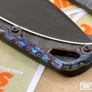 May include: Close-up of a custom knife with a dark blade and a handle featuring a vibrant, swirling pattern of blues, purples, and bronze hues. The handle is secured with two bolts. The knife is a Pacific Sons product.