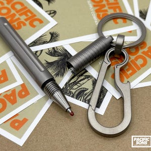 May include: A titanium keychain with a bottle opener, a carabiner, and a pen. The keychain is on top of several business cards with the text "Pacific Sons" printed on them.