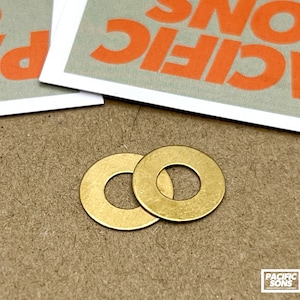 May include: Two brass washers with a central hole, displayed on a textured brown surface. The washers are circular and have a golden hue. The image also shows a card with the text "PACIFIC SONS" in orange.