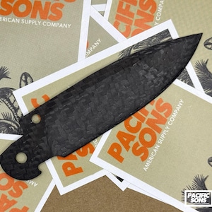 Puede incluir: Una hoja de cuchillo de fibra de carbono negra con un patrón de textura tejida y dos orificios circulares en el extremo de la espiga. La hoja está colocada sobre varias tarjetas rectangulares superpuestas con el texto Pacific Sons American Supply Company.