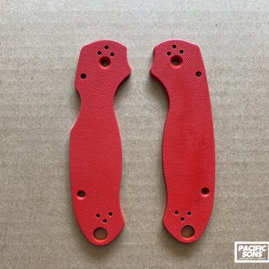 May include: Two red plastic knife scales with holes for mounting hardware. The scales are textured and have a slightly curved shape.