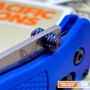 Può includere: Primo piano di un coltello pieghevole blu con una lama argentata. Il coltello presenta un'impugnatura blu strutturata e una vite di articolazione multicolore decorativa. La lama è contrassegnata dal logo "Benchmade".