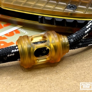 May include: Close-up of a clear amber-colored cord lock on a black and white braided cord. The cord lock has a cylindrical shape with a series of ridges and openings. The background includes a portion of a knife and a Pacific Sons label.